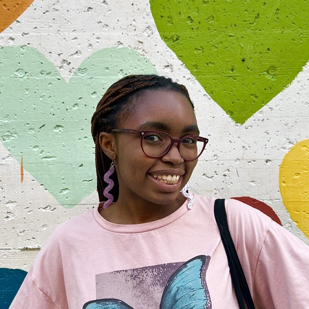 Headshot of a young Black woman with braids pulled back. She is smiling in front of a colorful wall.