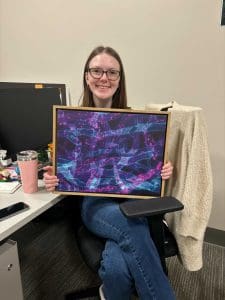 A young white woman smiling holding a framed canvas with a microscope image of fluorescent fungal hyphae and bacteria