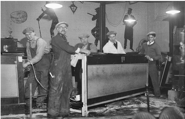 The agents pause yet again to pose for a photo amid the chaos of the ransacked speakeasy at 153 Causeway St. Prominently displayed in this photo are the caricatures painted on the wall of the hideaway. Two of the three figures are of high society men, both wearing top hats and one carrying a walking cane. The third figure wears a law officer's cap and carries a police baton, pointed at one of the other two figures, marking him as an agent of the law. He leans against one of the buildings support columns between the two other caricatures, an ironic reminder of the ever-present danger of being caught in such an establishment. The relaxed image of his leaning posture juxtaposed with his pointed baton denote the contrary legal status of alcohol sales at the time.
