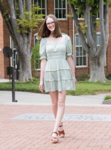 Full photo of a young, smiling white woman with glasses and light brown hair walking in a campus setting in a soft green dress.