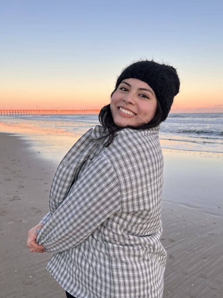 Headshot of a smiling, young latina woman in a checked coat and winter hat with a beach sunset in the background
