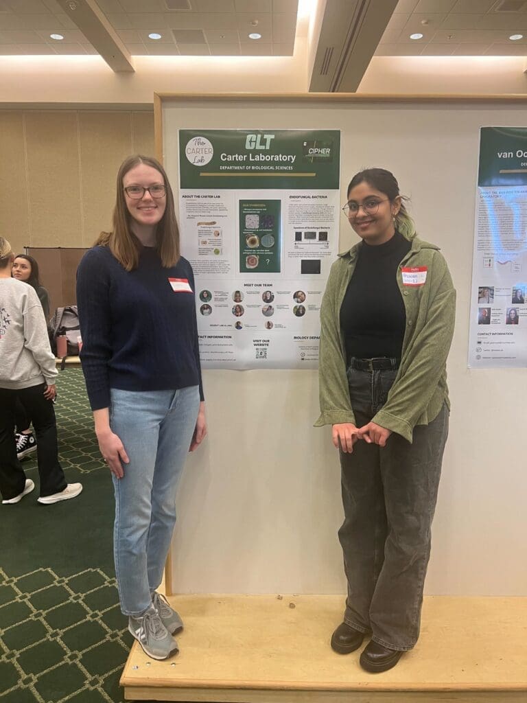 Two young women standing in front of a Carter Laboratory poster with images of fungi, the charlotte logo, and brief information about the lab.