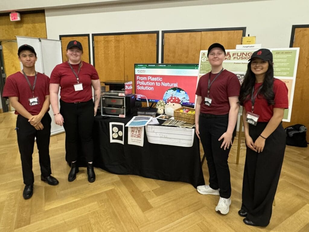 Four students in red tops, black pants, and black baseball caps stand in front of a table with various mushrooms and contraptions. The sign behind them says from plastic pollution to natural solution.
