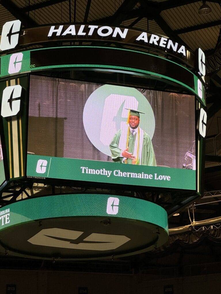 A Charlotte jumbotron screen showing a man in green graduation regalia with the name Timothy Chermaine Love listed underneath