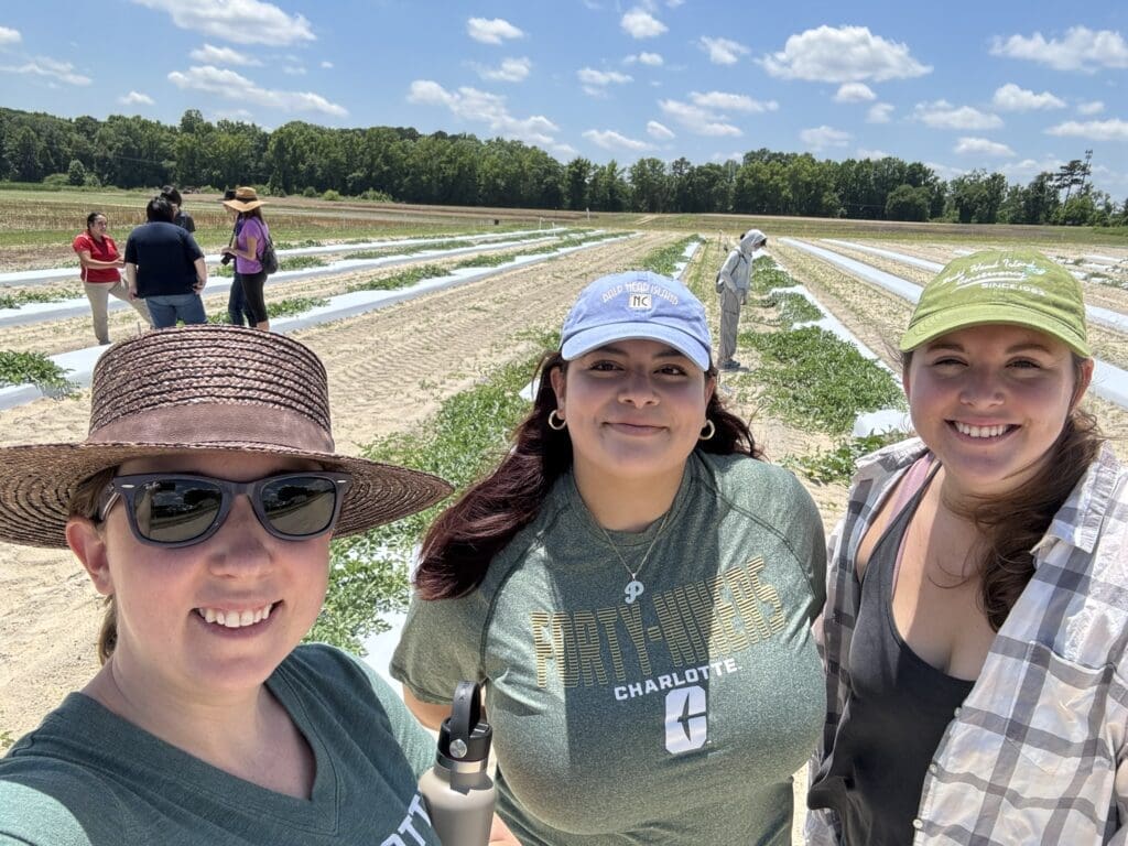 Three women in hats smiling for a selfie with farmland behind them.