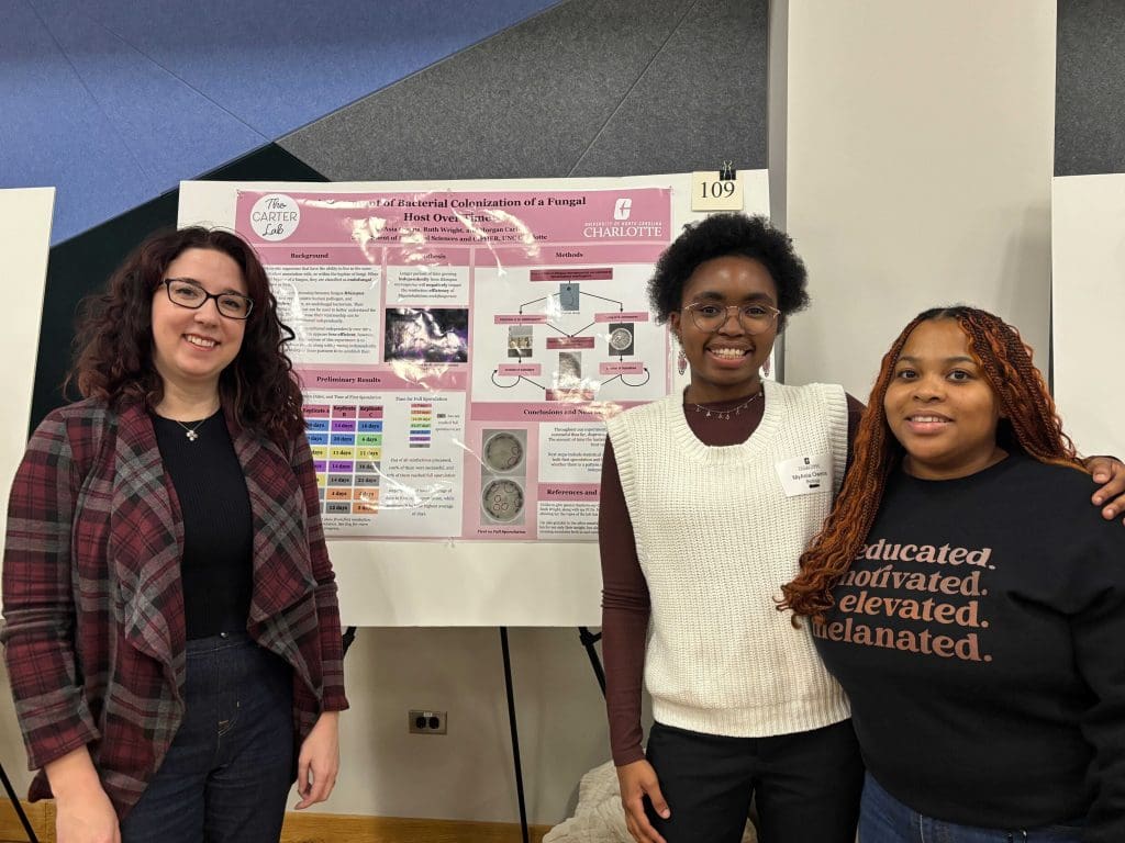 Three women smiling and standing in front of a pink scientific poster on bacterial-fungal interactions