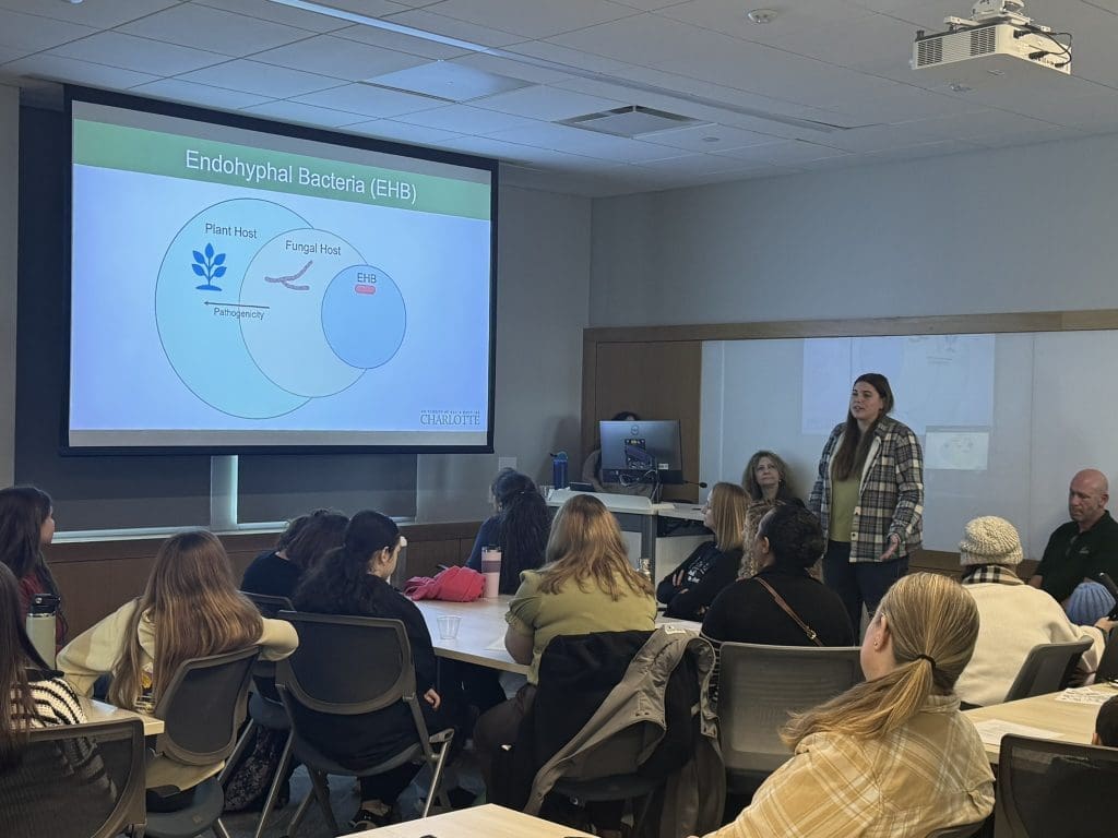 A woman giving a talk in front of a powerpoint screen on endohyphal bacteria and with many seated students looking on