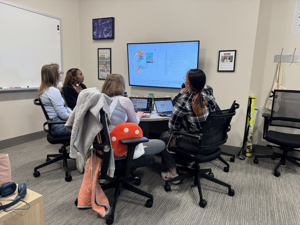 A group of four women clustered about a table looking up at a monitor with scientific-looking pictures on it of genome analyses